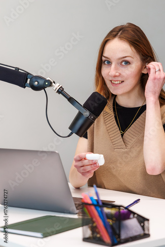 Fotomural A woman sits at a desk with a boom mic, adjusts an earbud, and holds its case