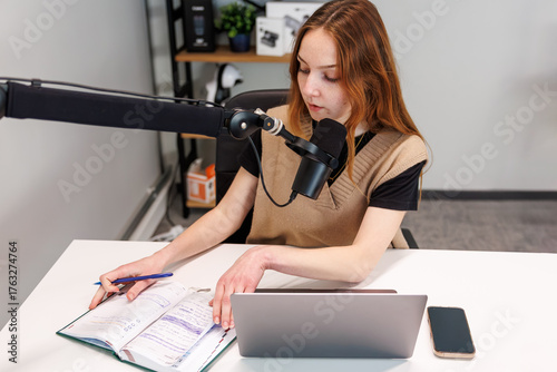 Cuadro en lienzo A young woman speaks into a boom mic, checks handwritten notes in a planner, and works beside a silver laptop and phone on a white desk in a small home studio