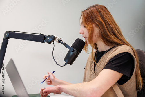 Fotomural A red haired woman speaks into a cardioid microphone on a boom arm, checks notes on a laptop, and holds a pen in an indoor studio with soft light and close side view