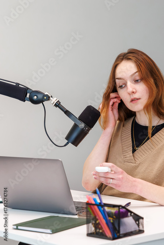 Cuadro en lienzo A young woman sits at a desk, adjusts an earbud, and speaks toward a boom arm microphone beside an open laptop, with soft light and tight framing in a studio setup