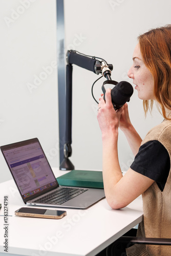 Fotomural A young woman speaks into a mounted condenser mic with pop filter, a laptop and smartphone on a white desk in a studio