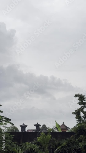 Dramatic cloudy sky with thick gray clouds forming above traditional temple roofs and tropical trees. Perfect atmospheric background for travel, religion, or nature themes in Southeast Asia.