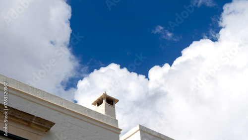Minimal white building with chimney under bright blue sky and fluffy white clouds, Mediterranean architecture style.