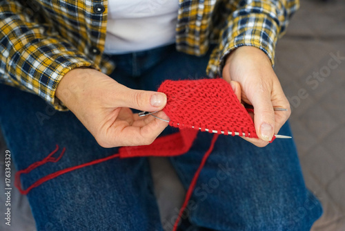 Woman in checkered shirt knitting red fabric with needles, close-up of hands and yarn. Cozy home hobby and creativity.