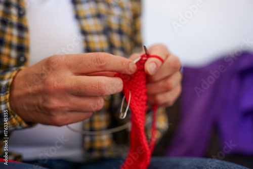 Woman in checkered shirt knitting red fabric with needles, close-up of hands and yarn. Cozy home hobby and creativity.