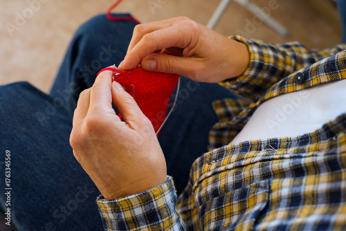 Woman in checkered shirt knitting red fabric with needles, close-up of hands and yarn. Cozy home hobby and creativity.