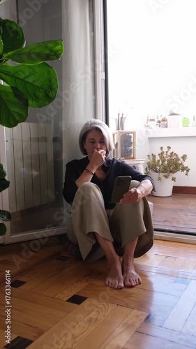 Mature woman using mobile phone at home, sitting barefoot on wooden floor by the balcony door