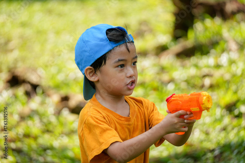 A cheerful young boy wearing an orange shirt and blue cap playing with a colorful water gun in a park