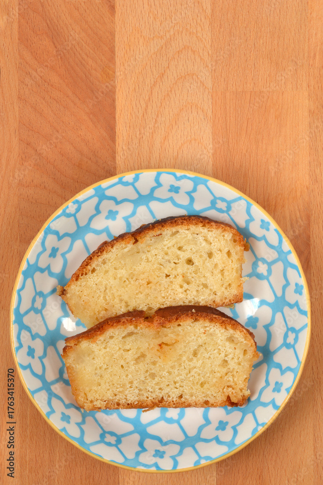 Obraz premium A vertical overhead shot featuring two slices of moist yellow cake with a golden crust, presented on a decorative blue and white plate on a light wooden surface