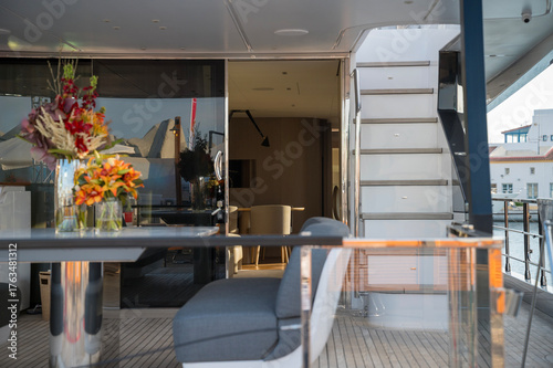 Elegant yacht interior with gray cushioned seating, modern table, glass railings, and fresh floral arrangements. Sunlight softly illuminates the cozy space, blending comfort with style.