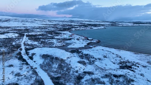 Aerial view of a remote, snow-covered arctic coastline featuring a small village or settlement with buildings and a pier by the large body of water