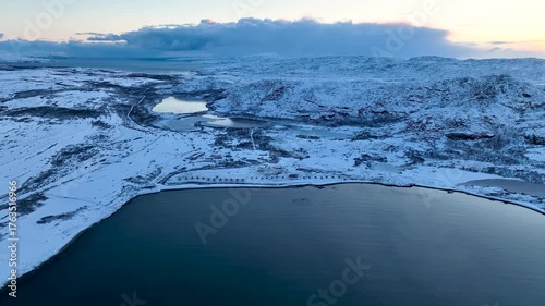 Aerial view of a remote arctic landscape with vast snow-covered hills, multiple frozen lakes, and a row of small domes or tents near the coastline and open water