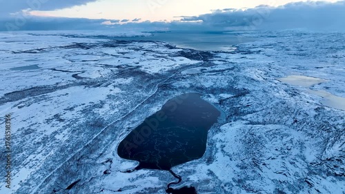 Aerial view of a vast, snow-covered arctic landscape with a large, dark, partially frozen lake in the foreground and a distant sea or bay under a cloudy sky