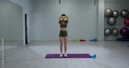 fit woman performs front dumbbell raises showcasing her strength and dedication to fitness while standing on a workout mat. Weights are visible nearby.