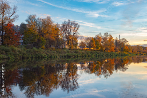 Calm autumn river reflecting colorful trees and houses under a blue sky during golden hour.