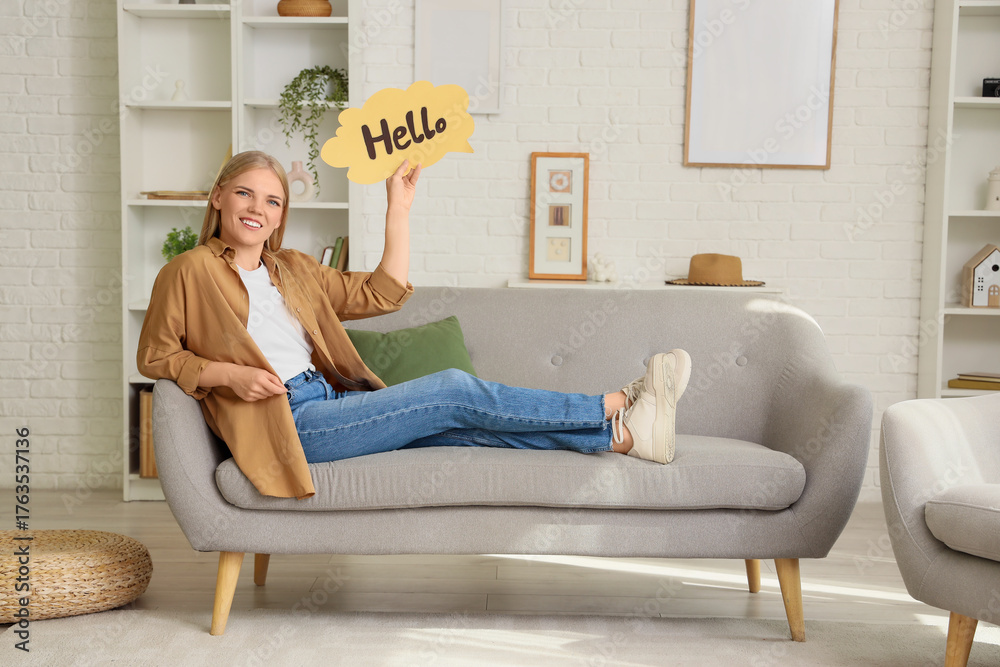 Obraz premium Young woman holding speech bubble with word HELLO on sofa at home