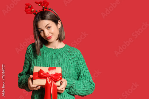 Young woman in reindeer horns with Christmas gift on red background
