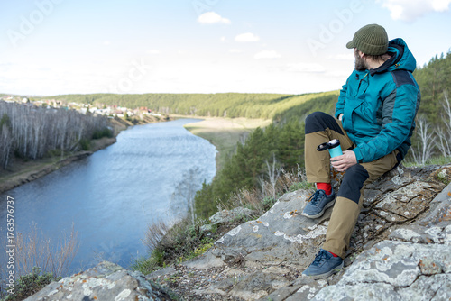 A bearded man is sitting resting on a rock with a thermocup in his hand, a knitted hat on his head