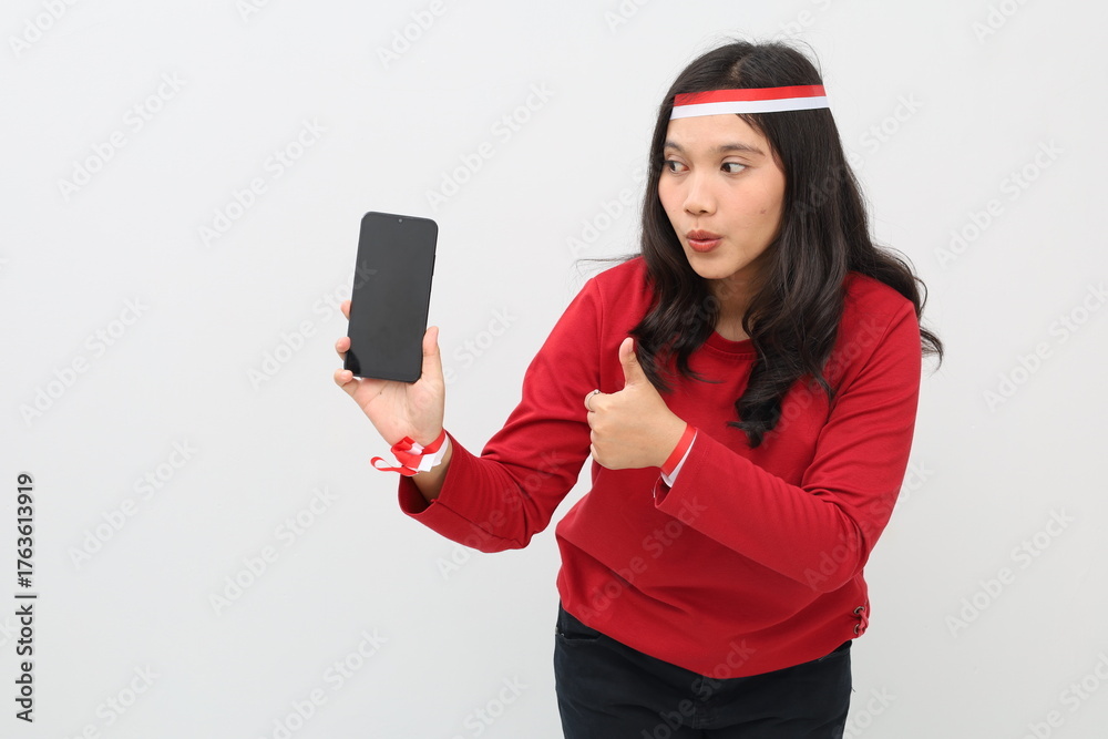 Naklejka premium Indonesian woman wearing red and white ribbon showing blank phone screen while giving thumbs up while looking at phone with funny facial expression, standing isolated on white background.