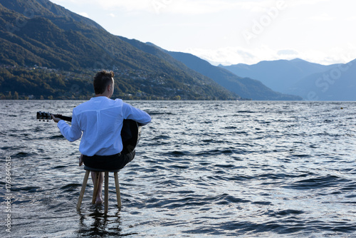 A young man sitting on a stool in the middle of a lake, seen from behind, playing the guitar under a slightly overcast sky.