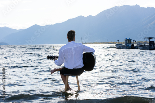 A young man sitting on a stool in the middle of a lake, seen from behind, playing the guitar under a slightly overcast sky.