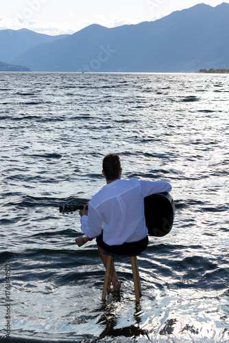 A young man sitting on a stool in the middle of a lake, seen from behind, playing the guitar under a slightly overcast sky.