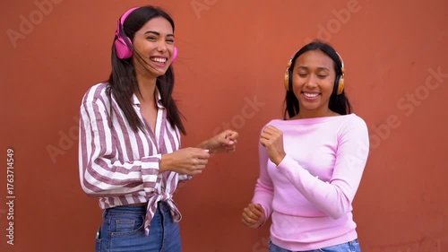 Happy young latin women dancing together in the city using wireless headphones - Technology, generation z and social media trend concept 
