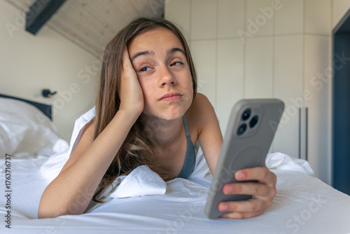 Girl bored with smartphone in white bed at morning. Sleepy doomscrolling.