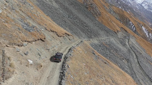 A vehicle skillfully navigates a rugged, winding dirt path through mountainous terrain, showcasing the natural landscape's textures and colors, with dynamic aerial camera movement enhancing the scene
