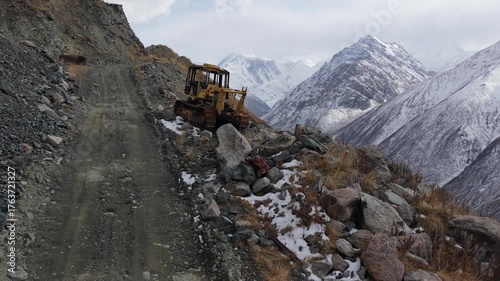 Heavy machinery is positioned on rocky terrain, gradually clearing large boulders while the camera zooms out to reveal the expansive mountainous landscape and rugged road conditions