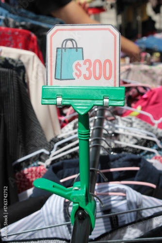 Placa de Promoção com Valor 300 em Feira de Moda Circular ao Ar Livre, Economia Circular e Roupas Second-Hand, Detalhe de Braço de Pessoa Selecionando Roupa ao Fundo