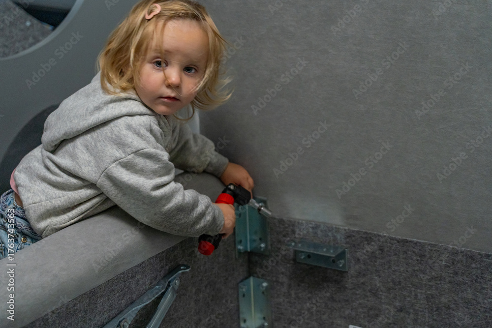 Naklejka premium Curious young blonde child intently participating in home improvement project, holding power drill and attempting to fasten metal brackets to gray surface.
