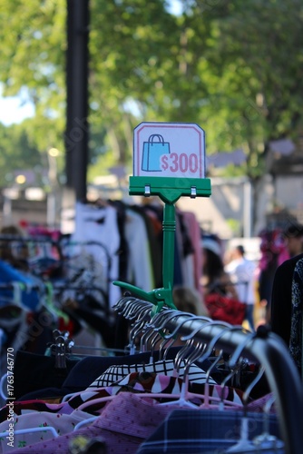 Feira de Moda Circular ao Ar Livre com Roupas em Araras, Clientes Desfocados ao Fundo e Placa de Promoção Destacando Valor 300, Ambiente Natural