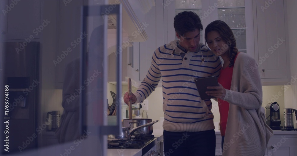 Fototapeta premium Cooking couple in hoodie, red top stirring stainless steel pot and checking tablet in home kitchen