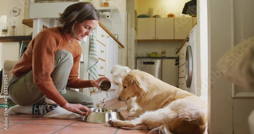 Photography Crouching woman pouring dog kibble from cup into steel bowl for dogs in kitchen,