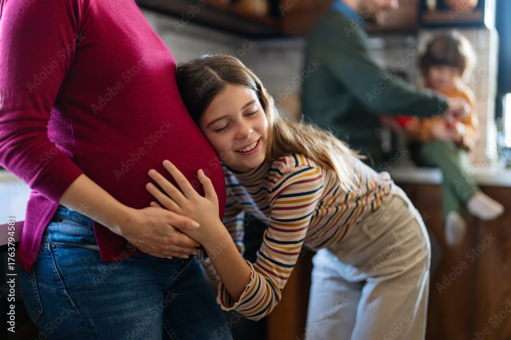 Fototapeta premium Happy daughter hugging belly her expecting mother at home. Excited girl listening to baby movement