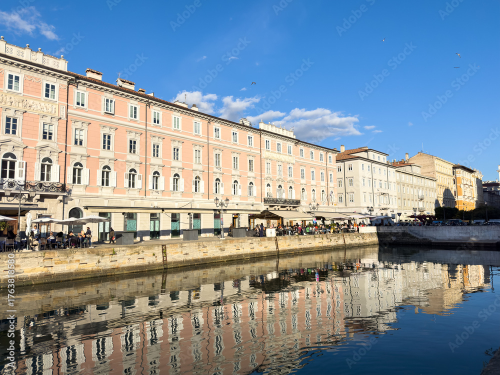 Fototapeta premium Trieste, Italy - October 14, 2025: Beautiful waterfront buildings reflect in calm water under clear blue sky in Trieste, Italy, showcasing stunning architecture and vibrant atmosphere