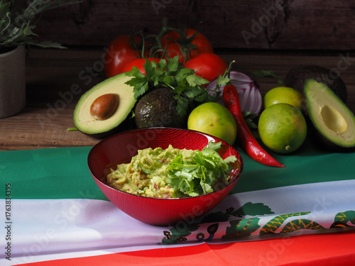 Guacamole in a red bowl, with ingredients at the back. All displayed over a Mexican flag.