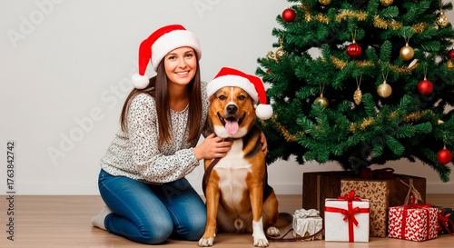 Festive fun with my furry friend! Woman and dog wear matching Santa hats by a Christmas tree filled with presents. Happy holidays!