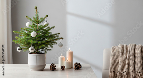 A miniature Christmas tree in a pot is ornamented with metallic baubles, illuminated candles, and pine cones on a table near a throw.