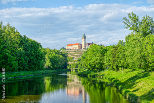 Landscape of Melnik with a calm rive with historic castle with a church tower. The scene is bright and peaceful, with clear skies and soft clouds adding to the charm of the view.