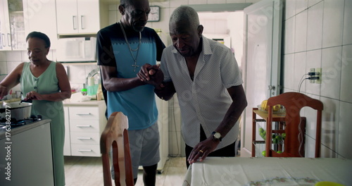 Elderly African American man being helped by his son to sit at a dining table in a cozy kitchen, reflecting care, compassion, and family connection in daily life