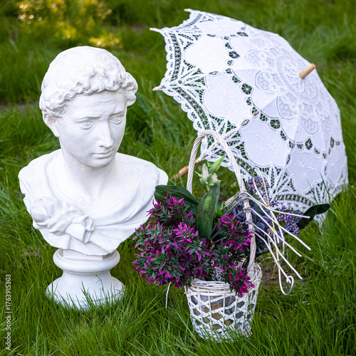 Elegant white classical bust, lace parasol, and wicker basket with purple flowers on lush green grass — romantic, vintage-inspired garden scene perfect for weddings, decor, or lifestyle themes.