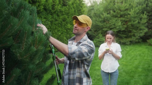 Man wearing protective glasses and a cap, actively pruning a conifer tree using long shears in a green garden, while a woman stands behind checking her smartphone