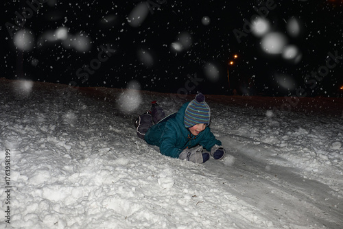 Child sliding down a snowy hill at night during snowfall, smiling and enjoying winter fun outdoors. Warm clothes and snow create a joyful holiday atmosphere