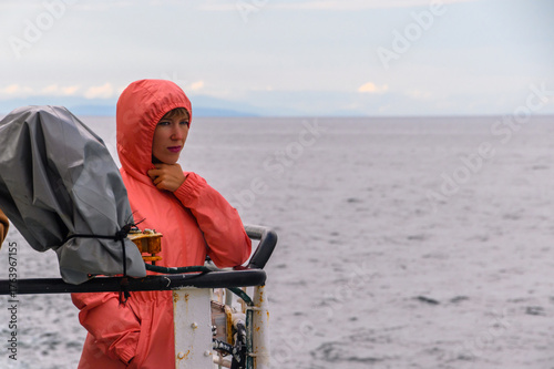 Woman in coral raincoat gazes at the ocean under overcast skies near a boat