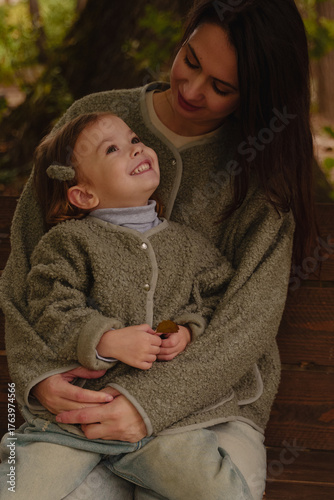 Mom hugs her daughter. Mom and daughter riding on a wooden swing in the park. A cheerful girl is sitting on her mother's lap. Warm gray jackets on mom and daughter. Mothers Day. Vertical photo.