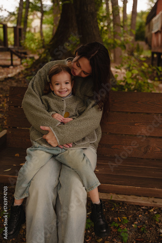 Mom and daughter riding on a wooden swing in the park. A cheerful girl is sitting on her mother's lap. Mom hugs her daughter. Warm gray jackets on mom and daughter. Vertical photo.