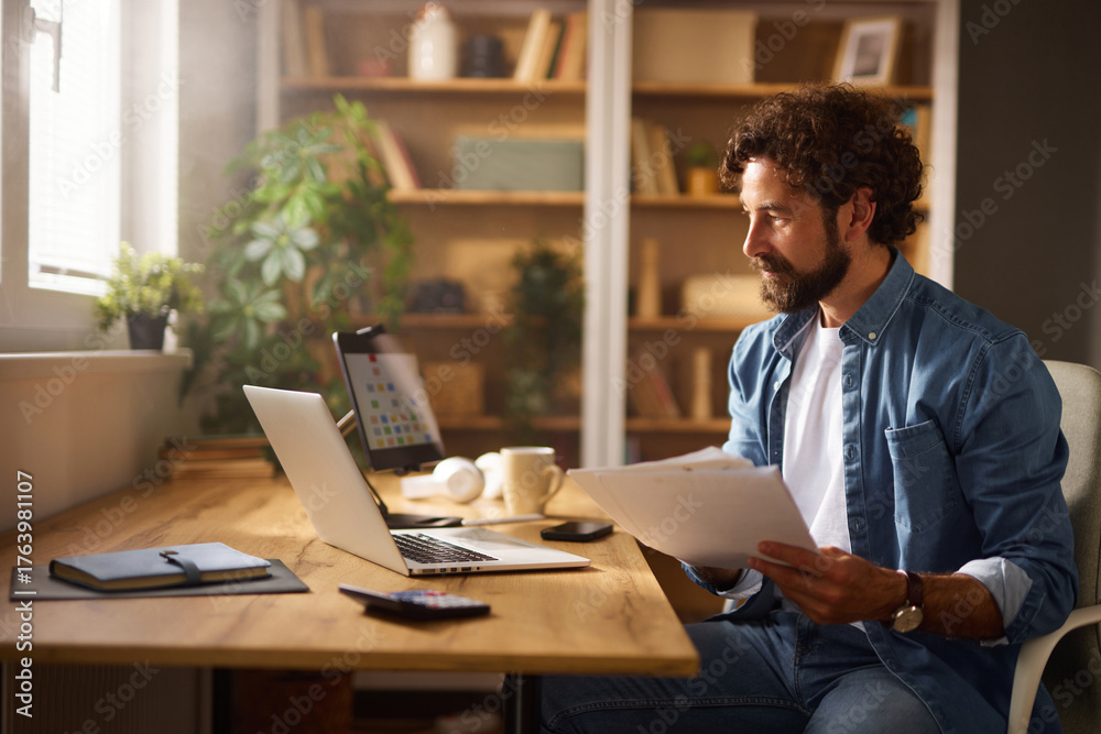 Obraz premium A man is engaged in work at a modern home office, seated at a wooden desk with a laptop and papers in front of him while surrounded by houseplants and shelves.