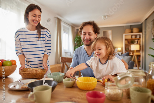 Obraz na plátně A joyful family dynamic unfolds as a mother lays out bowls and a father shares a playful moment with their child at a kitchen table